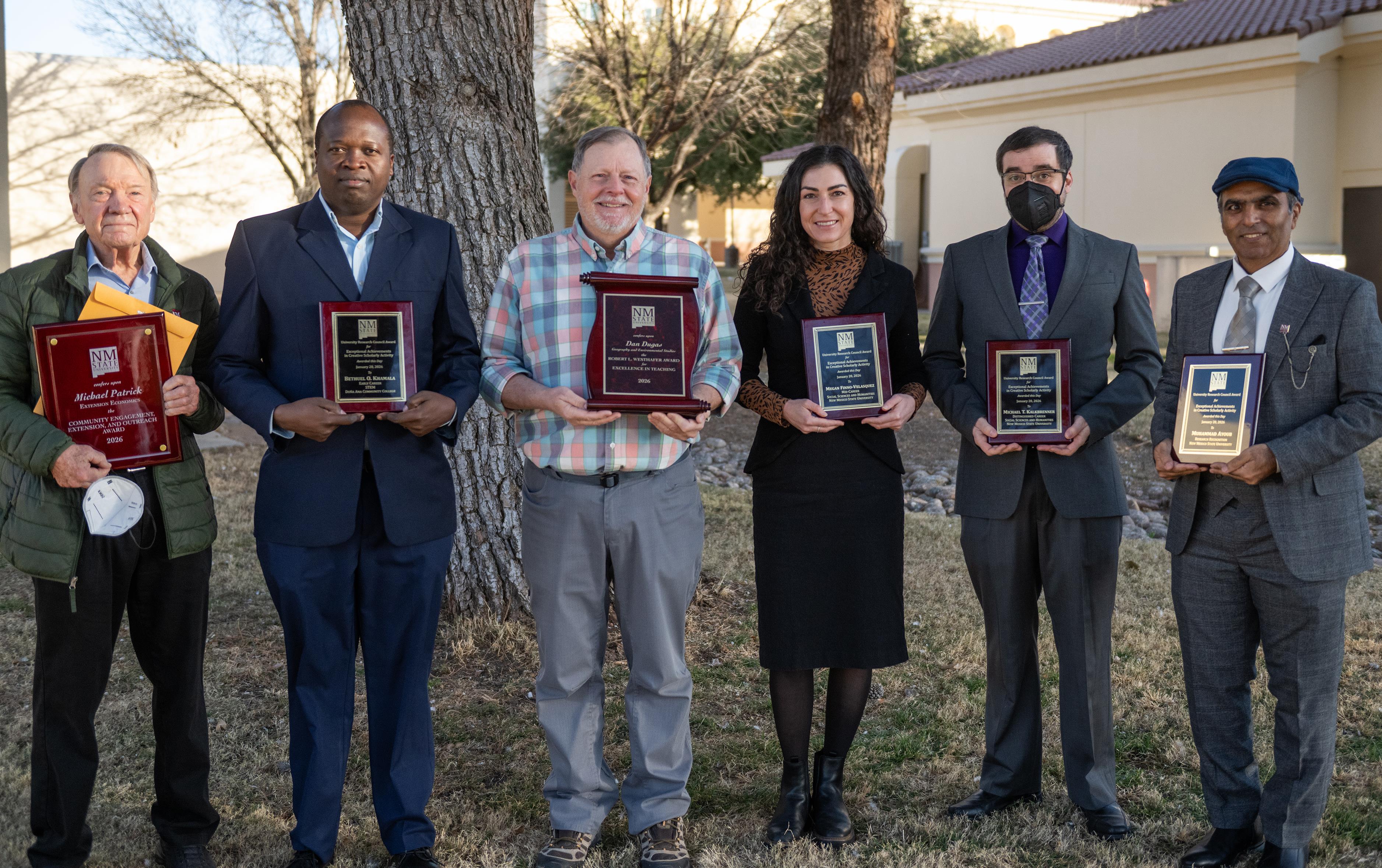 Group photo of six award recipients holding plaques in an outdoor setting.